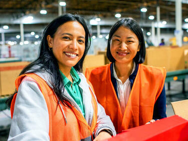 Twee vrouwen in oranje vesten die glimlachen in een warehouse